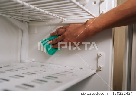 Close-up cropped shot of unrecognizable man defrosting and cleaning interior of refrigerator using blue rag to wipe away ice and grime buildup, ensuring fresh and sanitary food storage environment. Close-up cropped shot of unrecognizable man defrosting and cleaning interior of refrigerator using blue rag to wipe away ice and grime buildup, ensuring fresh and sanitary food storage environment. 138222424