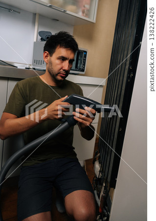Vertical portrait of focused homeowner using vacuum cleaner to remove dust and debris from refrigerator coils, performing essential maintenance for optimal cooling efficiency and energy saving Vertical portrait of focused homeowner using vacuum cleaner to remove dust and debris from refrigerator coils, performing essential maintenance for optimal cooling efficiency and energy saving 138222426