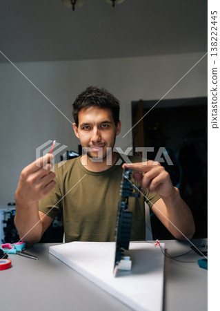 Vertical portrait of skilled computer service technician repairing computer part while holding cotton swab and looking at camera, sitting at desk in workshop. Concept of service, cleaning computer. Vertical portrait of skilled computer service technician repairing computer part while holding cotton swab and looking at camera, sitting at desk in workshop. Concept of service, cleaning computer. 138222445