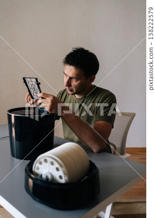 Vertical portrait of focused man holding inspecting air humidifier filter, performing maintenance and cleaning to ensure clean, healthy air in home environment. Concept of maintenance home appliance. 138222579
