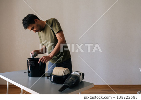 Portrait of young man disassembling and cleaning air humidifier filter standing by table in living room, performing regular maintenance for healthy air quality. Concept of household cleanliness. Portrait of young man disassembling and cleaning air humidifier filter standing by table in living room, performing regular maintenance for healthy air quality. Concept of household cleanliness. 138222583