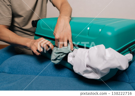 Close-up cropped shot of traveler struggling with an overpacked turquoise suitcase, compressing clothes to zip it for an upcoming trip or vacation, facing packing challenges with excessive luggage. Close-up cropped shot of traveler struggling with an overpacked turquoise suitcase, compressing clothes to zip it for an upcoming trip or vacation, facing packing challenges with excessive luggage. 138222716