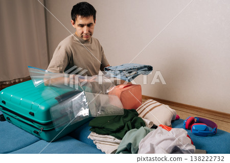 Puzzled young man packing various garments and essentials into a large suitcase with clear vacuum bags, maximizing space for an upcoming journey, trip, or move. Puzzled young man packing various garments and essentials into a large suitcase with clear vacuum bags, maximizing space for an upcoming journey, trip, or move. 138222732