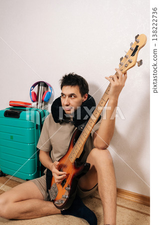 Portrait of young man in travel neck pillow sits by a turquoise suitcase in an airport terminal, playing bass guitar and wearing headphones relaxed, ready for a musical journey and travel adventure. Portrait of young man in travel neck pillow sits by a turquoise suitcase in an airport terminal, playing bass guitar and wearing headphones relaxed, ready for a musical journey and travel adventure. 138222736