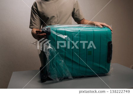 Cropped shot of backpacker male preparing luggage for travel, protecting large suitcase with clear plastic wrap, ensuring items are safe before departure from home. Cropped shot of backpacker male preparing luggage for travel, protecting large suitcase with clear plastic wrap, ensuring items are safe before departure from home. 138222749