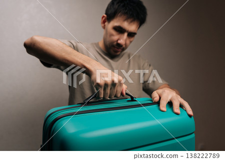 Young man traveler standing indoors, pulling up the retractable handle of a teal hard-shell suitcase as he prepares and packs for an imminent trip or weekend getaway. Young man traveler standing indoors, pulling up the retractable handle of a teal hard-shell suitcase as he prepares and packs for an imminent trip or weekend getaway. 138222789