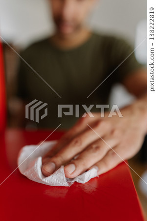 Vertical cropped shot of unrecognizable man wiping dry red surface of shelf with white paper towel after housekeeping, close-up. Concept of cleanliness and hygiene in domestic environment. Vertical cropped shot of unrecognizable man wiping dry red surface of shelf with white paper towel after housekeeping, close-up. Concept of cleanliness and hygiene in domestic environment. 138222819