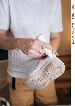 Vertical cropped shot of unrecognizable person using bottle of shoe whitener and painting scuffed surface of dirty white sneaker, illustrating process of cleaning, recoloring, and restoring footwear. Vertical cropped shot of unrecognizable person using bottle of shoe whitener and painting scuffed surface of dirty white sneaker, illustrating process of cleaning, recoloring, and restoring footwear. 138222861