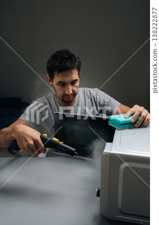 Portrait of man cleaning microwave oven with steam cleaner and sponge, using hot vapor for powerful disinfection and removing dirt, representing effective household chore and modern cleaning solution. 138222877
