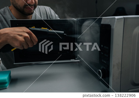 Close-up of man cleaning and sanitizing microwave oven with steam cleaner device, eliminating germs and tough stains, maintaining hygiene in kitchen at home, dark room. 138222906