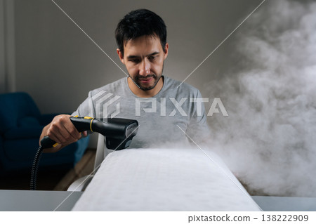 Portrait of young adult man using handheld steam cleaner to sanitize and refresh pillow through chemical-free deep cleaning, ensuring clean and hygienic sleeping space and preventing allergens. Portrait of young adult man using handheld steam cleaner to sanitize and refresh pillow through chemical-free deep cleaning, ensuring clean and hygienic sleeping space and preventing allergens. 138222909