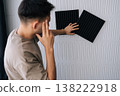 Rear view of pensive man placing square black acoustic foam panels on a textured white soundproof wall, creating an effective barrier for soundproofing and room acoustics. 138222918