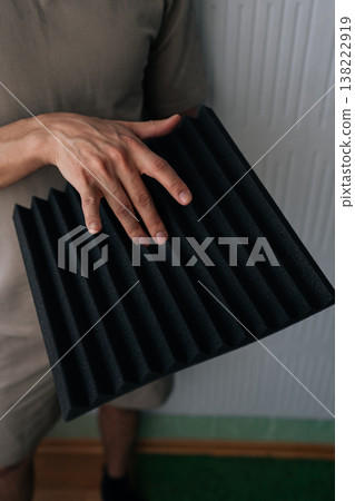 Close-up hand of handyman holding a black acoustic foam panel, exploring soundproofing materials for studio environment, noise reduction, and audio quality improvement projects. 138222919