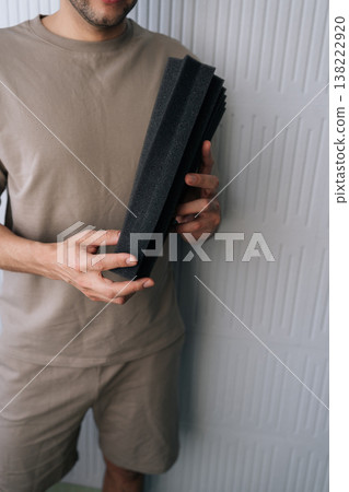 Man holding multiple black acoustic foam panels, preparing for the soundproofing process by applying insulation to a wall for recording studio or home audio setup. 138222920