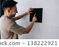 Smiling man installing black acoustic foam panel on white decorative wall to reduce echo, improve sound quality in a home studio or recording space during renovation project. 138222921