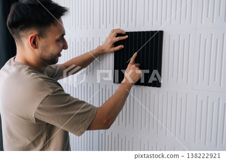 Smiling man installing black acoustic foam panel on white decorative wall to reduce echo, improve sound quality in a home studio or recording space during renovation project. Smiling man installing black acoustic foam panel on white decorative wall to reduce echo, improve sound quality in a home studio or recording space during renovation project. 138222921
