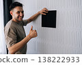 Portrait of cheerful handyman man installing a black acoustic panel on a soundproof wall, creating a modern interior design for home sound insulation and noise reduction from reverberation. 138222933