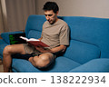 Skilled developer siting on blue sofa studies textbook while laptop shows green code lines, illustrating focused remote learning, skill development and modern software study at home. 138222934