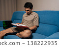 Focused programmer male siting on blue sofa studies textbook while laptop shows green code lines, illustrating focused remote learning, skill development and modern software study at home. 138222935