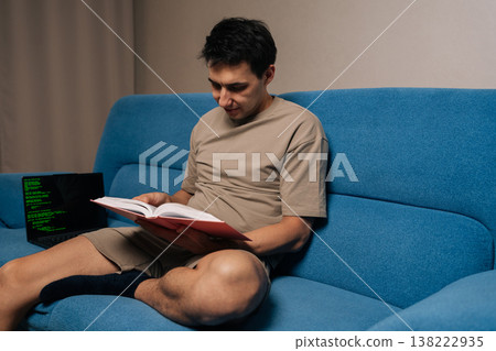 Focused programmer male siting on blue sofa studies textbook while laptop shows green code lines, illustrating focused remote learning, skill development and modern software study at home. Focused programmer male siting on blue sofa studies textbook while laptop shows green code lines, illustrating focused remote learning, skill development and modern software study at home. 138222935
