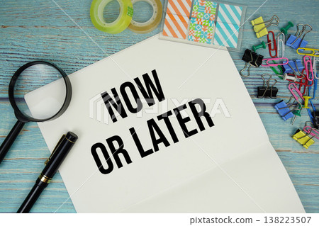 Now Or Later text on a paper surrounded by office supplies and magnifying glass on a blue wooden surface 138223507