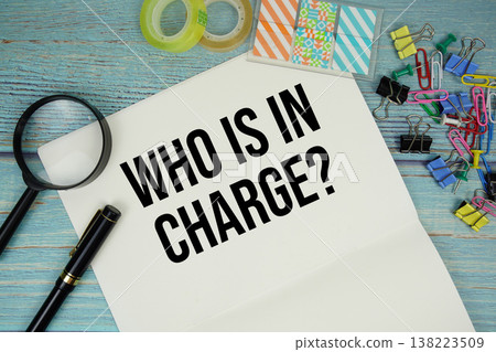 Who Is In Charge? text on a paper surrounded by office supplies and magnifying glass on a blue wooden surface Who Is In Charge? text on a paper surrounded by office supplies and magnifying glass on a blue wooden surface 138223509