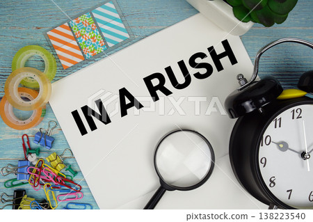 In A Rush text on a paper surrounded by office supplies, a magnifying glass, and a clock on a blue wooden surface In A Rush text on a paper surrounded by office supplies, a magnifying glass, and a clock on a blue wooden surface 138223540