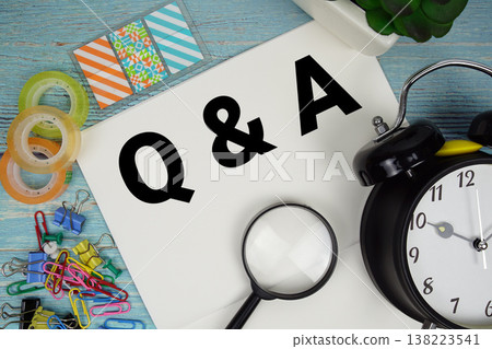 Q and A (Question and Answer) text on a paper surrounded by office supplies, a magnifying glass, and a clock on a blue wooden surface 138223541