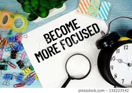 Become More Focused text on a paper surrounded by office supplies, a magnifying glass, and a clock on a blue wooden surface 138223542