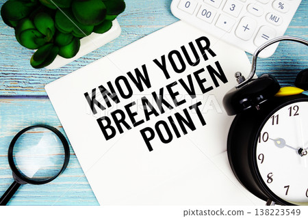 Know Your Breakeven Point message written on paper placed on a blue wooden surface, alongside a magnifying glass, a clock, a calculator, and a small plant 138223549
