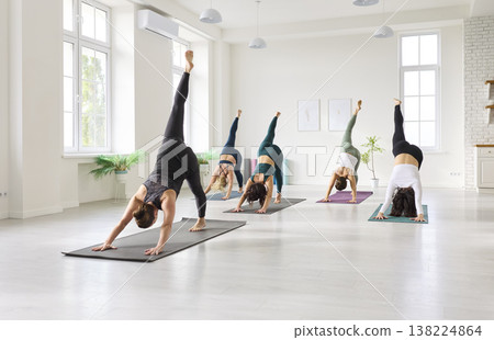 Yoga class lifting legs in three-legged dog pose inside a bright studio interior 138224864
