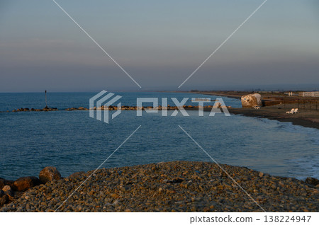 Serene coastal landscape at dusk with gentle waves lapping against rocky shore and distant silhouettes Serene coastal landscape at dusk with gentle waves lapping against rocky shore and distant silhouettes 138224947