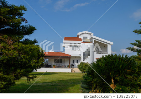 Elegant white villa surrounded by lush greenery under a bright blue sky in a serene landscape Elegant white villa surrounded by lush greenery under a bright blue sky in a serene landscape 138225075