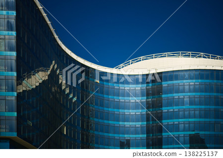 Modern Glass Building with Curved Facade and Blue Tinted Windows, Contemporary Corporate Architecture Reflecting Clear Blue Sky, Urban Business District Exterior Detail. 138225137