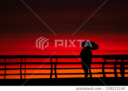 Silhouette of a person with an umbrella against a fiery red sunset. A minimalist high-contrast shot of a figure on a pier railing set against an intense orange and crimson evening sky 138225140