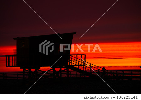 Silhouette of a lifeguard tower against a dramatic red sunset. A high-contrast shot of a coastal structure on stilts with stairs, set against a vibrant orange and dark crimson evening sky. Silhouette of a lifeguard tower against a dramatic red sunset. A high-contrast shot of a coastal structure on stilts with stairs, set against a vibrant orange and dark crimson evening sky. 138225141