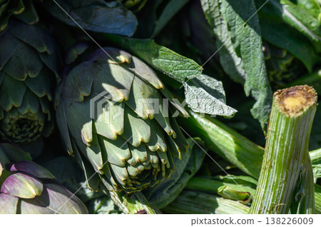 Fresh artichokes nestled among vibrant greens at a bustling farmers market under bright sunlight 138226009