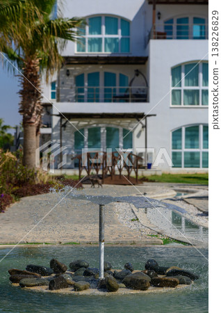 Close-up of water jets from a fountain creating a refreshing splash in a pool, with droplets catching the sunlight and forming ripples on the surface. 138226829