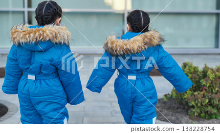 Little sisters walking outdoors in matching blue winter jumpsuits with fur hoods 138228754