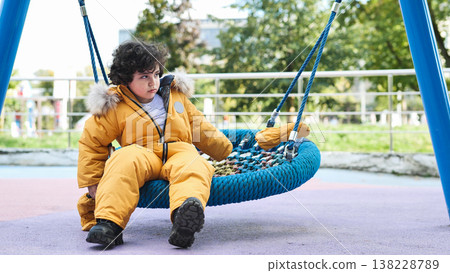 Thoughtful boy wearing warm yellow winter jumpsuit sitting on playground swing Thoughtful boy wearing warm yellow winter jumpsuit sitting on playground swing 138228789