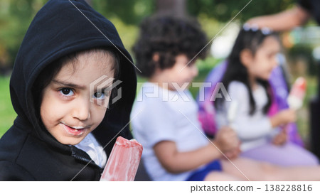 Young child eating popsicle looking at camera with friends in a summer park Young child eating popsicle looking at camera with friends in a summer park 138228816