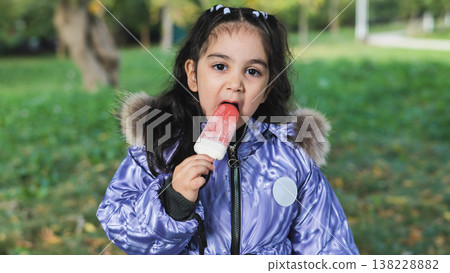 Little girl enjoying a sweet popsicle on a cool autumn day in a green park 138228882