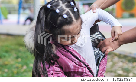 Caring parent helping little girl put on her warm pink jacket in a park Caring parent helping little girl put on her warm pink jacket in a park 138228884