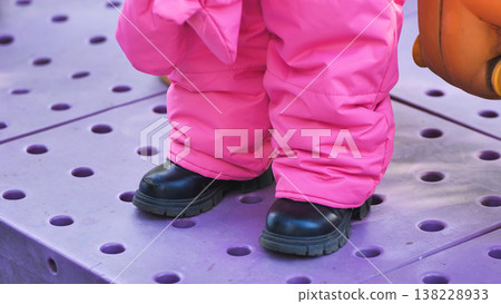 Child wearing pink snowsuit and winter boots standing on purple playground surface Child wearing pink snowsuit and winter boots standing on purple playground surface 138228933