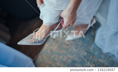 Close up of bride's hands fastening elegant white high heeled shoes with crystals, preparing for wedding ceremony Close up of bride's hands fastening elegant white high heeled shoes with crystals, preparing for wedding ceremony 138229087