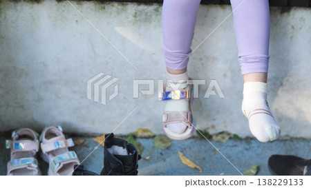 Child's changing footwear, wearing one sandal and one sock, other shoes on ground Child's changing footwear, wearing one sandal and one sock, other shoes on ground 138229133