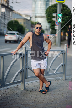 Muscular man wearing sunglasses, shorts and a tank top is leaning on a fence on a city street in russia Muscular man wearing sunglasses, shorts and a tank top is leaning on a fence on a city street in russia 138229216