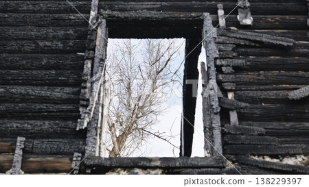Burnt facade of a wooden house with a window opening showing a bare tree and sky Burnt facade of a wooden house with a window opening showing a bare tree and sky 138229397