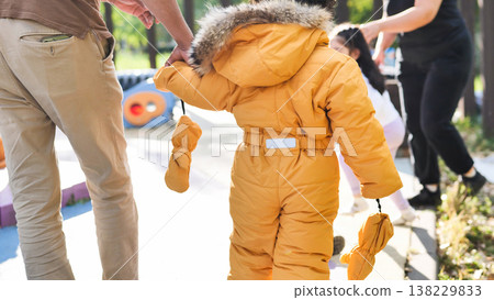 Parent holding toddler hand, walking together on playground in yellow snowsuit 138229833