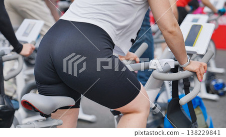 Rear view of a woman in sportswear working out on a stationary bike during a group fitness class. Rear view of a woman in sportswear working out on a stationary bike during a group fitness class. 138229841